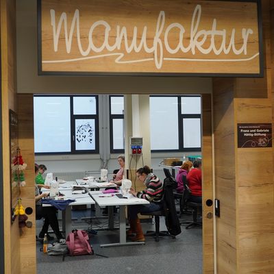 Blick durch die Tür in die offene Manufaktur. Dort sitzen Mitarbeiter aus den Werkstätten an Nähmaschinen.